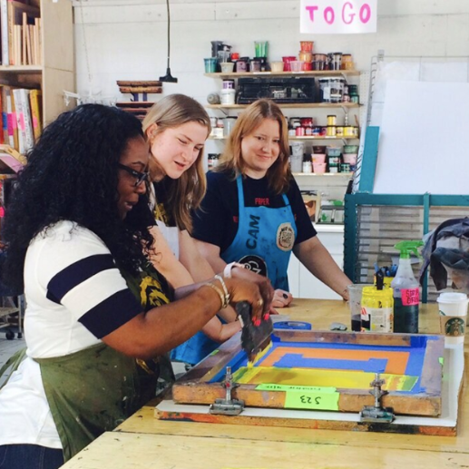Three people screen print on paper using a print board and squeegee.