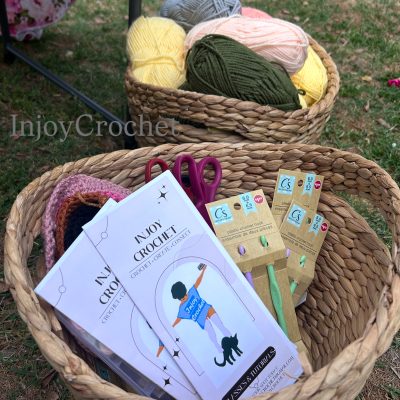 2 Baskets - lower basket holding crochet needles, scissors, and flyers with company logo and “InjoyCrochet”. Upper basket is filled with yarn (yellow, green, pink, grey). 