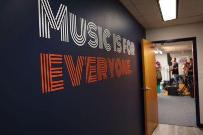 Alt Text:  Sound Hall Studios hallway wall featuring the phrase "Music Is for Everyone" in bold white and orange retro typography on a navy background, with a rehearsal studio visible in the background with musicians playing.